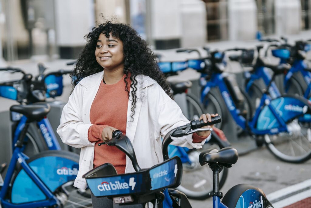 Cheerful African American female bicyclist in casual clothes renting bicycle in bicycle sharing station on city street and looking away contentedly