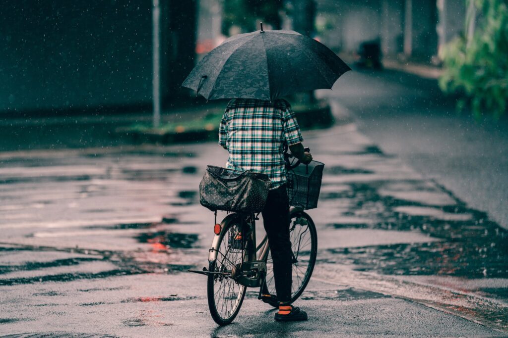 Person in Plaid Shirt and Black Pants Holding an Umbrella While Riding a Bicycle